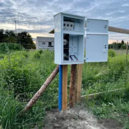 Otwarta, biała skrzynka elektryczna zamontowana na drewnianych słupkach, z niebieską rurą osłonową na przewody, na tle łąki i zachmurzonego nieba.
