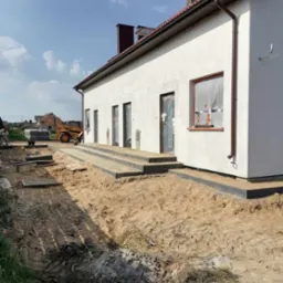 Budynek jednorodzinny w trakcie prac wykończeniowych elewacji, z widocznymi schodami wejściowymi, zabezpieczonymi oknami i przygotowanym terenem wokół, na którym stoi żółta koparko-ładowarka.
