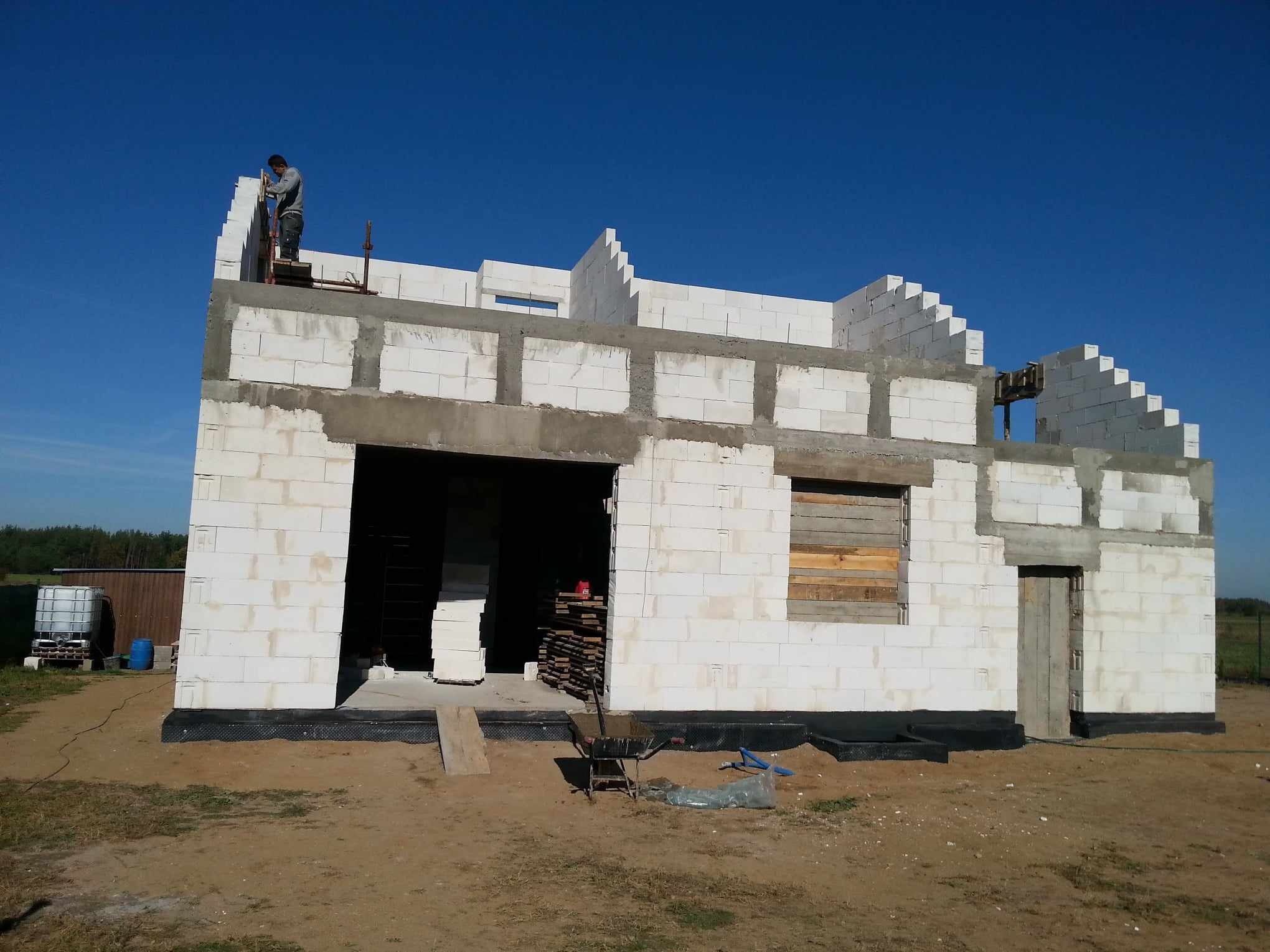 Budowa domu z białych bloczków, widoczny murarz pracujący na rusztowaniu, teren budowy z materiałami i narzędziami, okno zabite deskami.