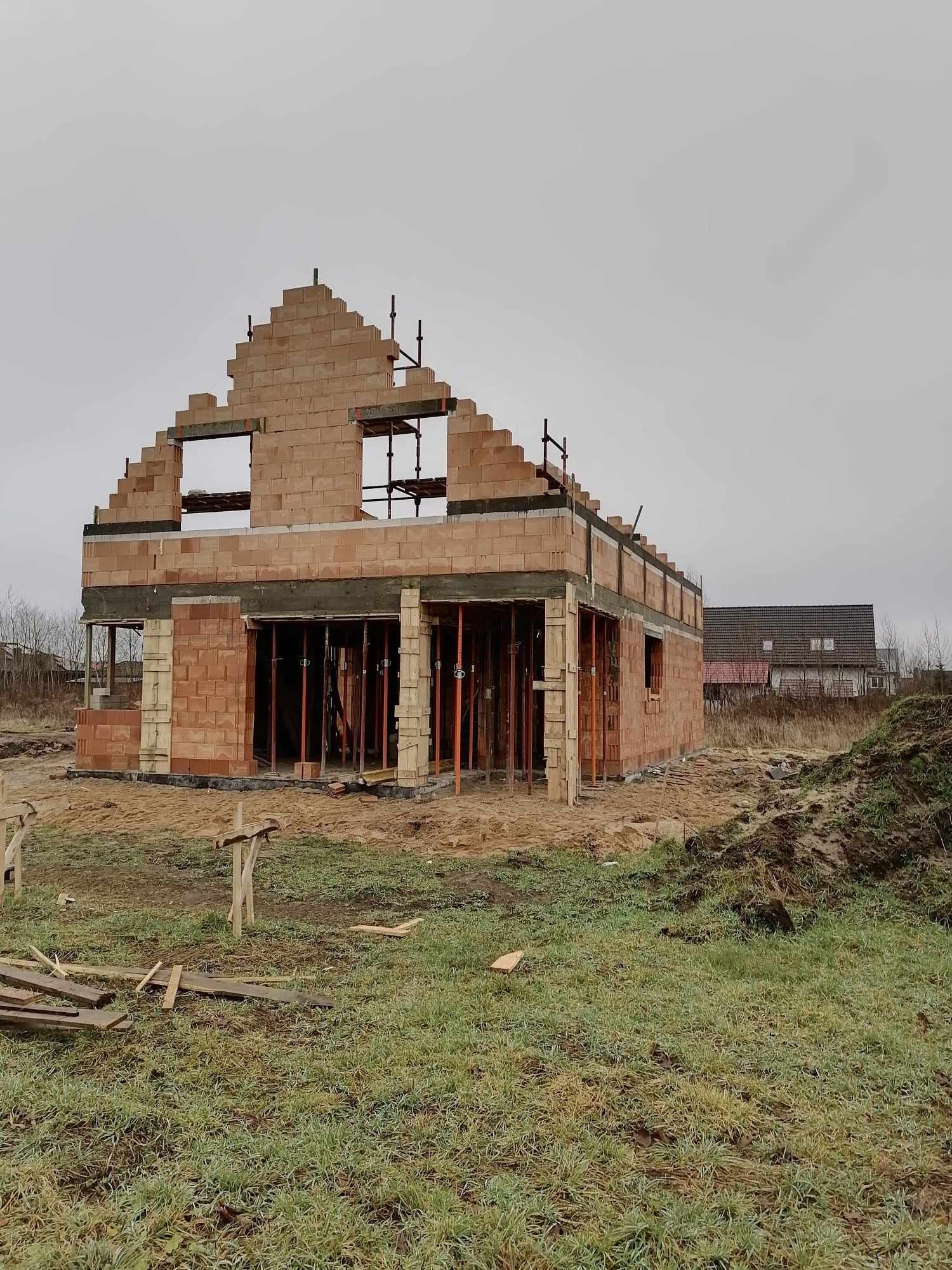 Budowa domu jednorodzinnego w trakcie wznoszenia ścian z bloczków, widoczne rusztowania i podpory, pochmurne niebo w tle.