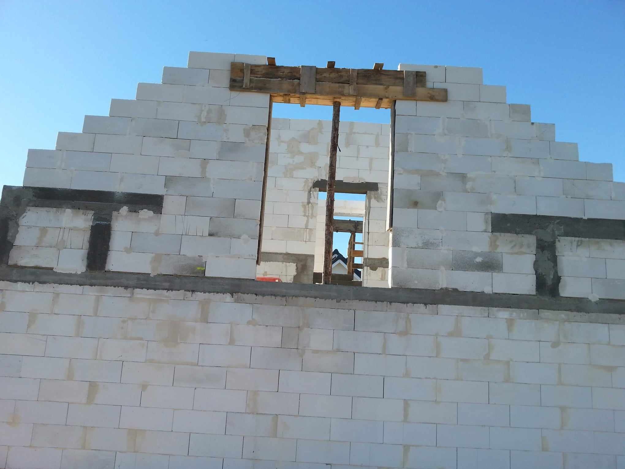 Wznoszenie ściany z bloczków białych betonu komórkowego, widoczne drewniane szalunki okienne i fragment konstrukcji domu w tle.