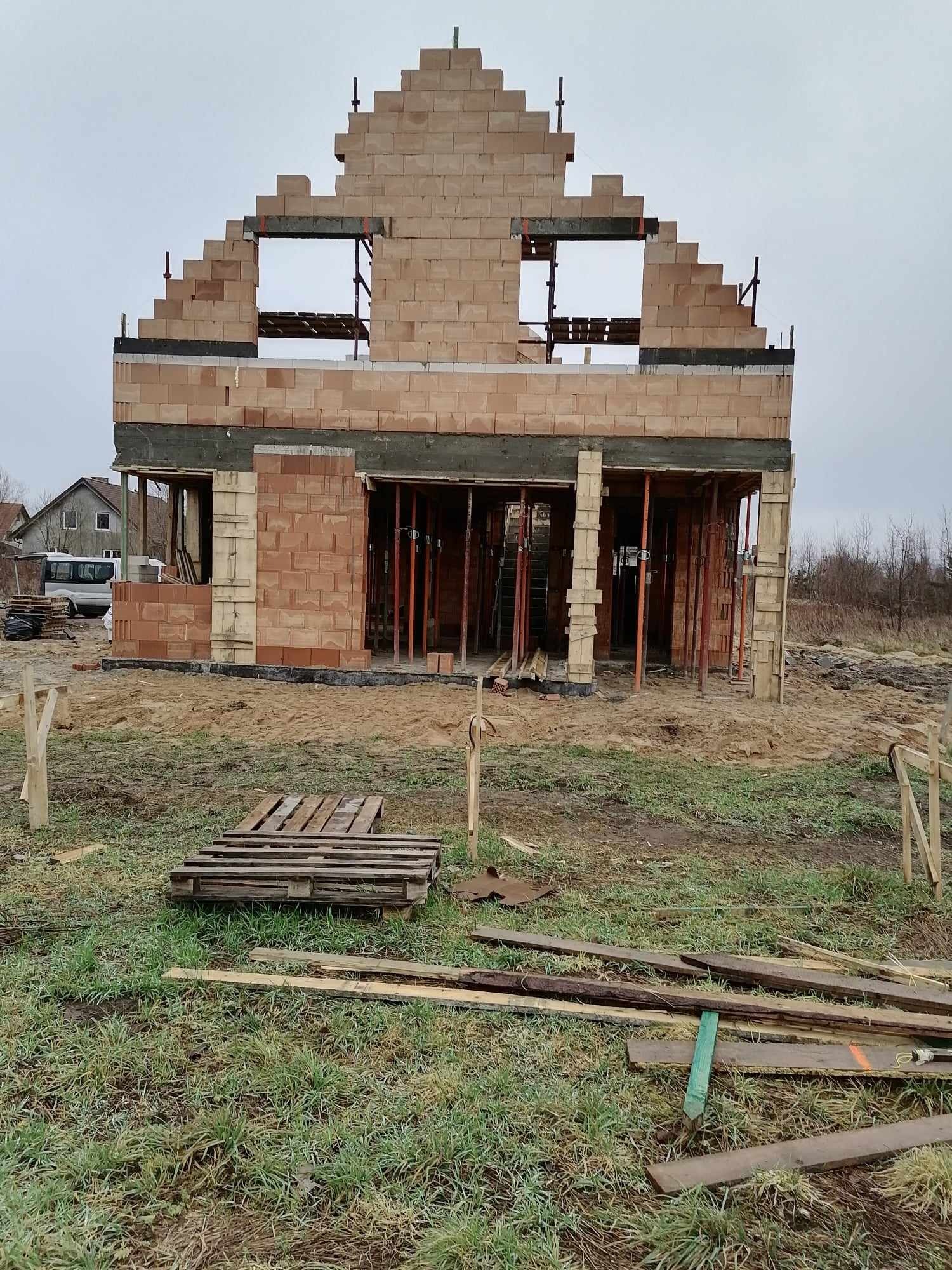 Wznoszenie dwukondygnacyjnego budynku z pustaków ceramicznych; widoczne otwory okienne i elementy szalunkowe, na pierwszym planie paleta i deski na trawiastym terenie.