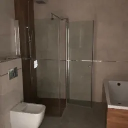 Nowoczesna łazienka z kabiną prysznicową typu walk-in, podwieszanym bidetem i zabudowaną wanną z drewnianym akcentem, wykończona w szarościach i drewnie.