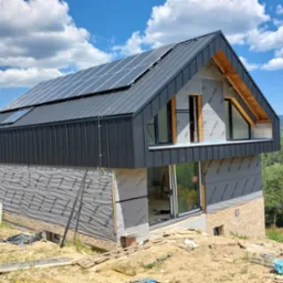 Nowoczesny dom w trakcie budowy z zamontowanym dachem z blachy na rąbek stojący i panelami fotowoltaicznymi, widoczne okna dachowe i membrana na ścianach.