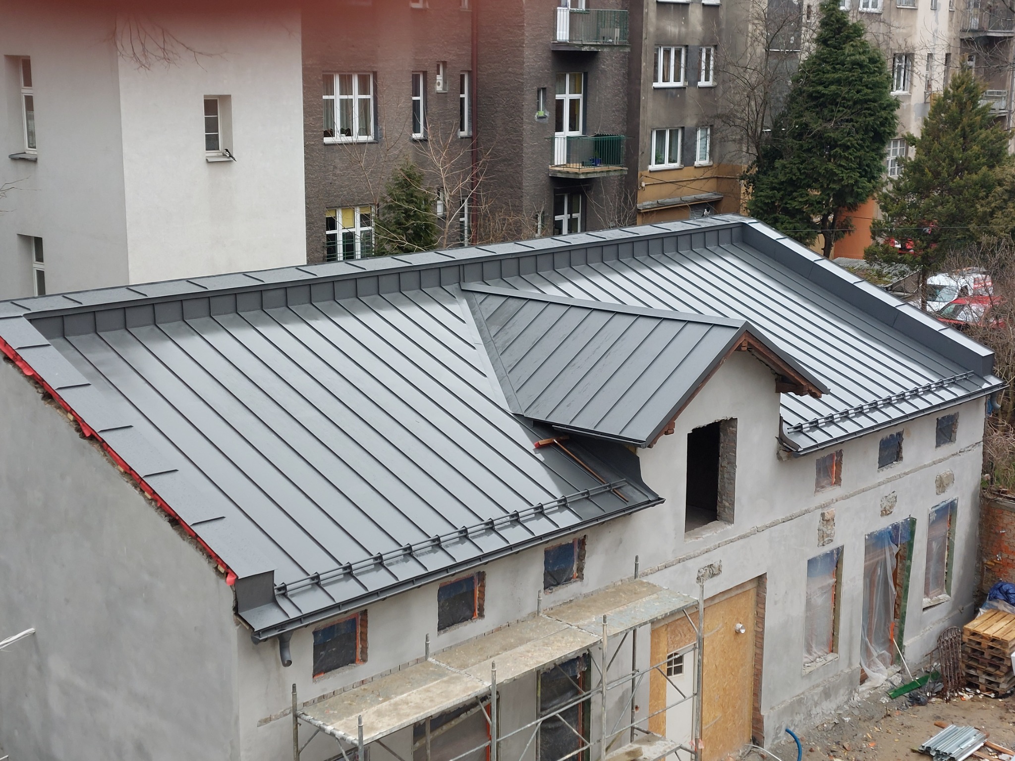 Nowy, szary dach z blachy na budynku w trakcie remontu, widoczne rusztowania i zabezpieczone okna folią.