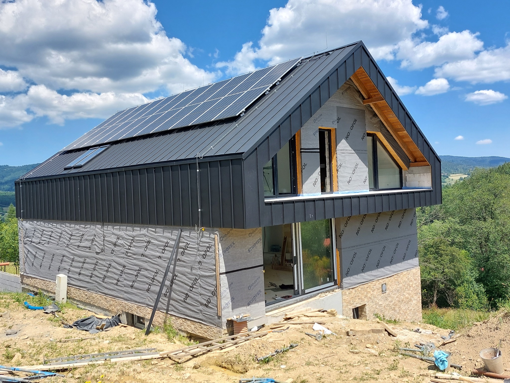 Nowoczesny dom w trakcie budowy z zamontowanym dachem z blachy na rąbek stojący i panelami fotowoltaicznymi, widoczne okna dachowe i membrana na ścianach.