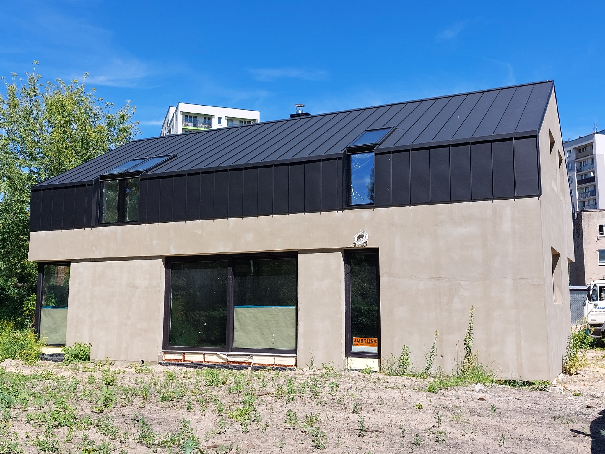 Nowoczesny, dwukondygnacyjny dom z ciemną, metalową konstrukcją dachu, lukarnami i surowym, jasnym tynkiem elewacyjnym, otoczony niezagospodarowanym terenem z widoczną roślinnością.