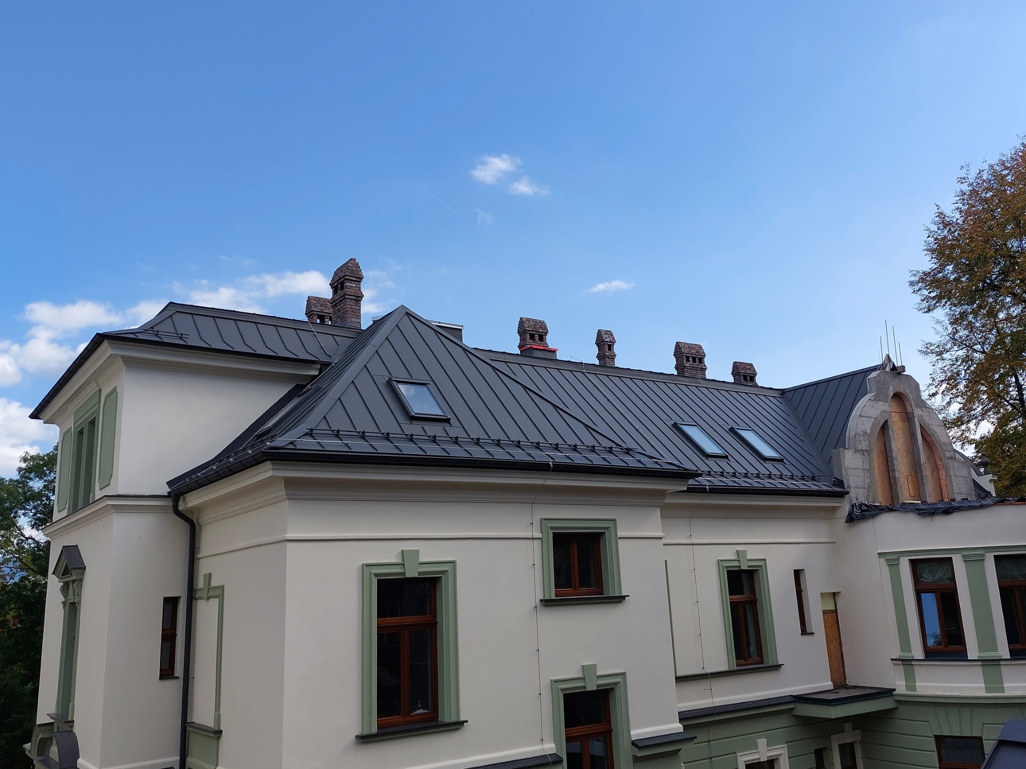 Odnowiony budynek z ciemnym metalowym dachem, widoczne kominy z cegły i okna dachowe, na tle błękitnego nieba z lekkimi chmurami.
