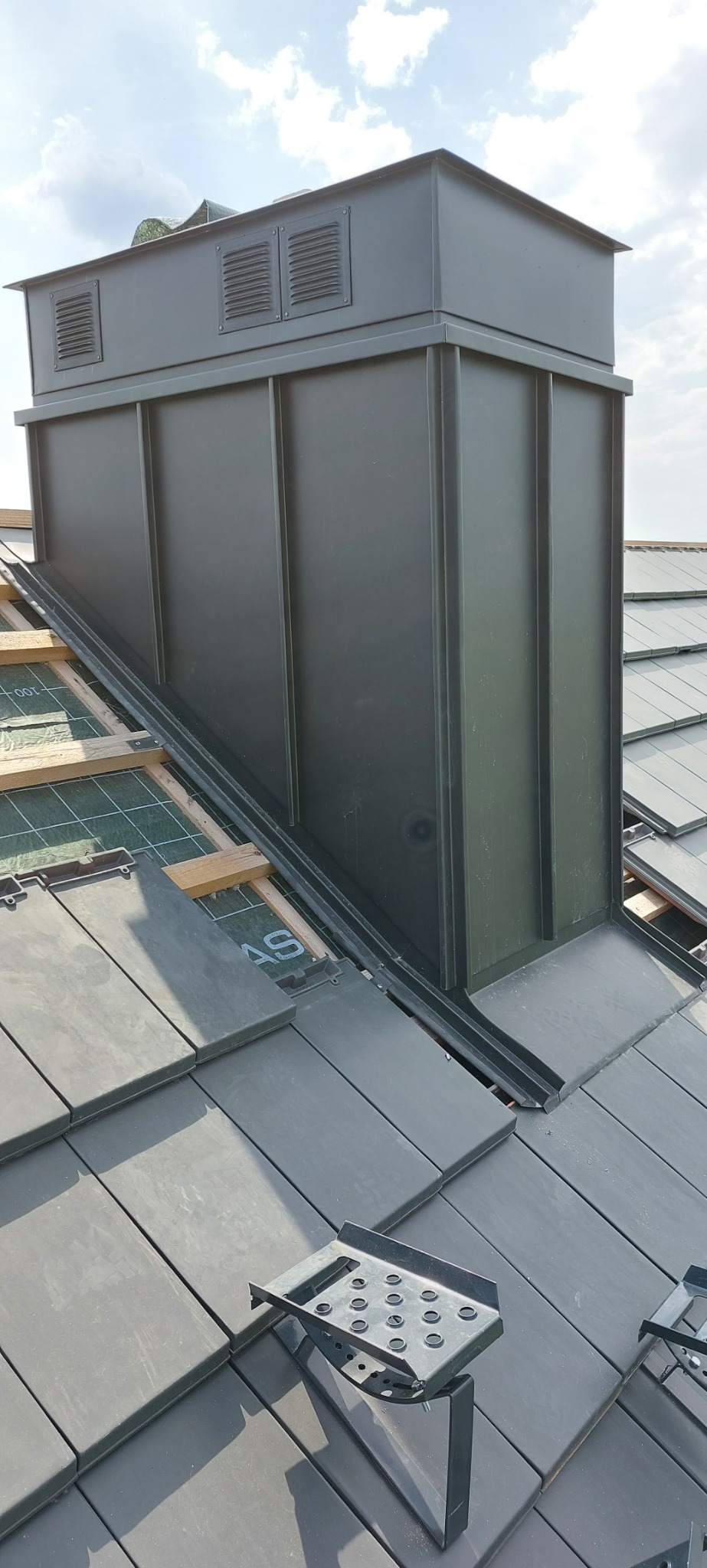 Nowoczesny, grafitowy komin wentylacyjny z blachy na dachu z dachówki ceramicznej, widoczne elementy konstrukcji dachu i uchwyt monterski