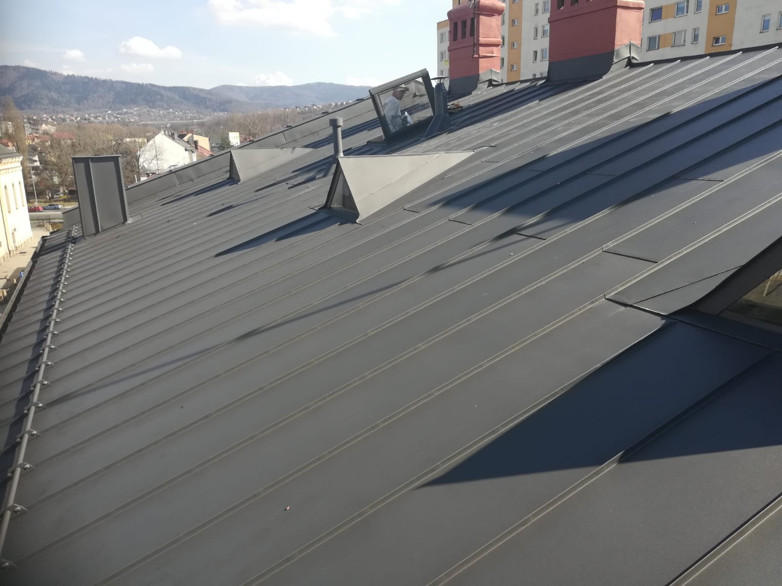 Nowoczesny, grafitowy dach z blachy trapezowej z oknami dachowymi i kominami wentylacyjnymi na tle gór i zabudowy miejskiej.