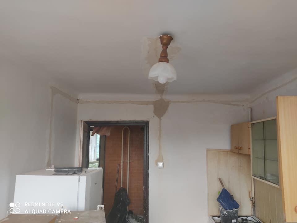 Wnętrze pokoju w trakcie odświeżania ścian, widoczne ślady po szpachlowaniu i malowaniu, wisząca lampa z drewnianym elementem, fragmenty mebli kuchennych i lodówki.