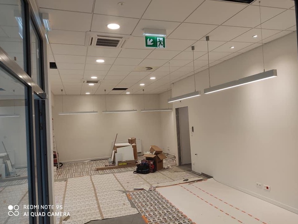 Wnętrze w trakcie remontu: podłoga z ogrzewaniem podłogowym w trakcie instalacji, widoczne kable i maty grzewcze, białe ściany, sufit podwieszany z oświetleniem liniowym, kartony i materiały...