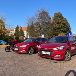 El-Mot Jan cempiel - Dwa czerwone samochody z oznaczeniem nauki jazdy na placu manewrowym, obok motocykl z kursantem w kasku i czerwonej kurtce, w tle budynek z czerwonym dachem i drzewa jesienią.