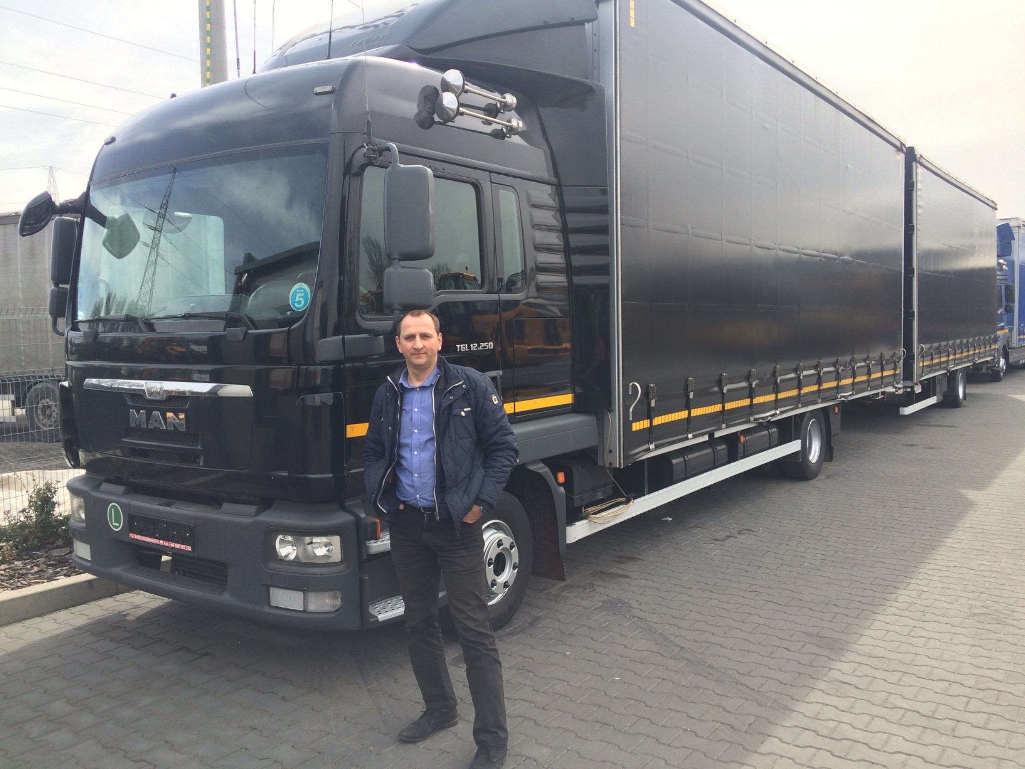 Czarny ciągnik siodłowy MAN TGL 12.250 z naczepą kurtynową i mężczyzna w kurtce na tle kostki brukowej.