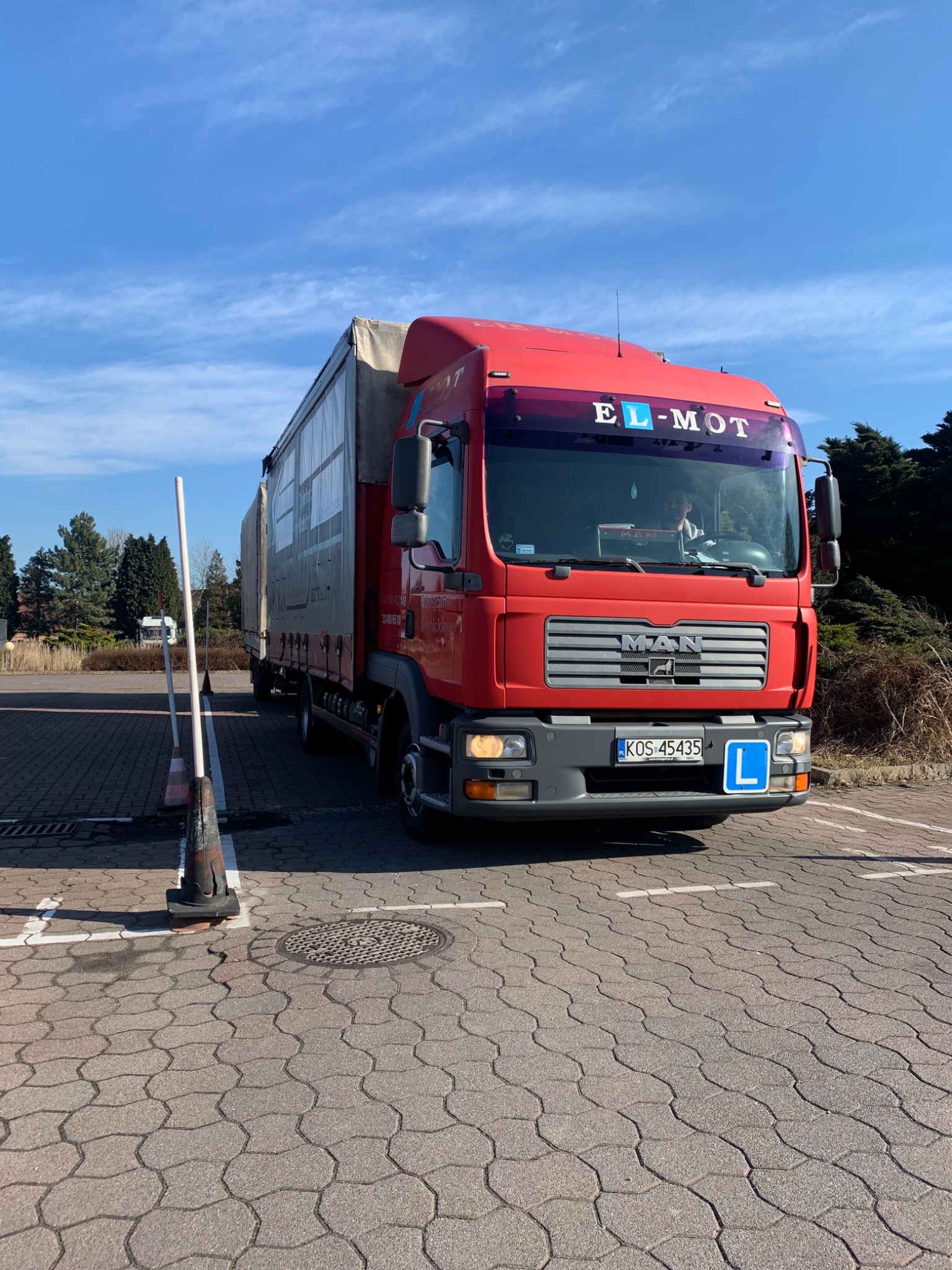 Czerwona ciężarówka MAN z oznaczeniem 'L' (nauka jazdy) na parkingu, widoczne tablice rejestracyjne KOS 45435 i logo firmy EL-MOT nad przednią szybą.