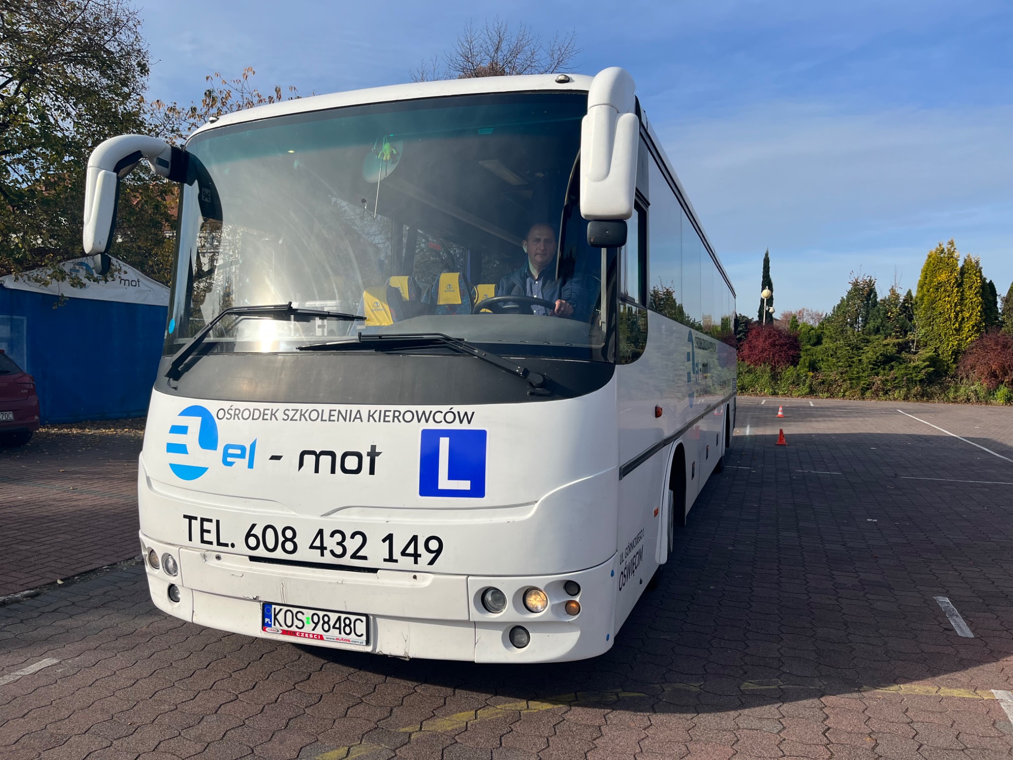 Biały autobus z napisem 'Ośrodek Szkolenia Kierowców el-mot' i niebieską literą 'L' na parkingu, widok z przodu na kierowcę za szybą.