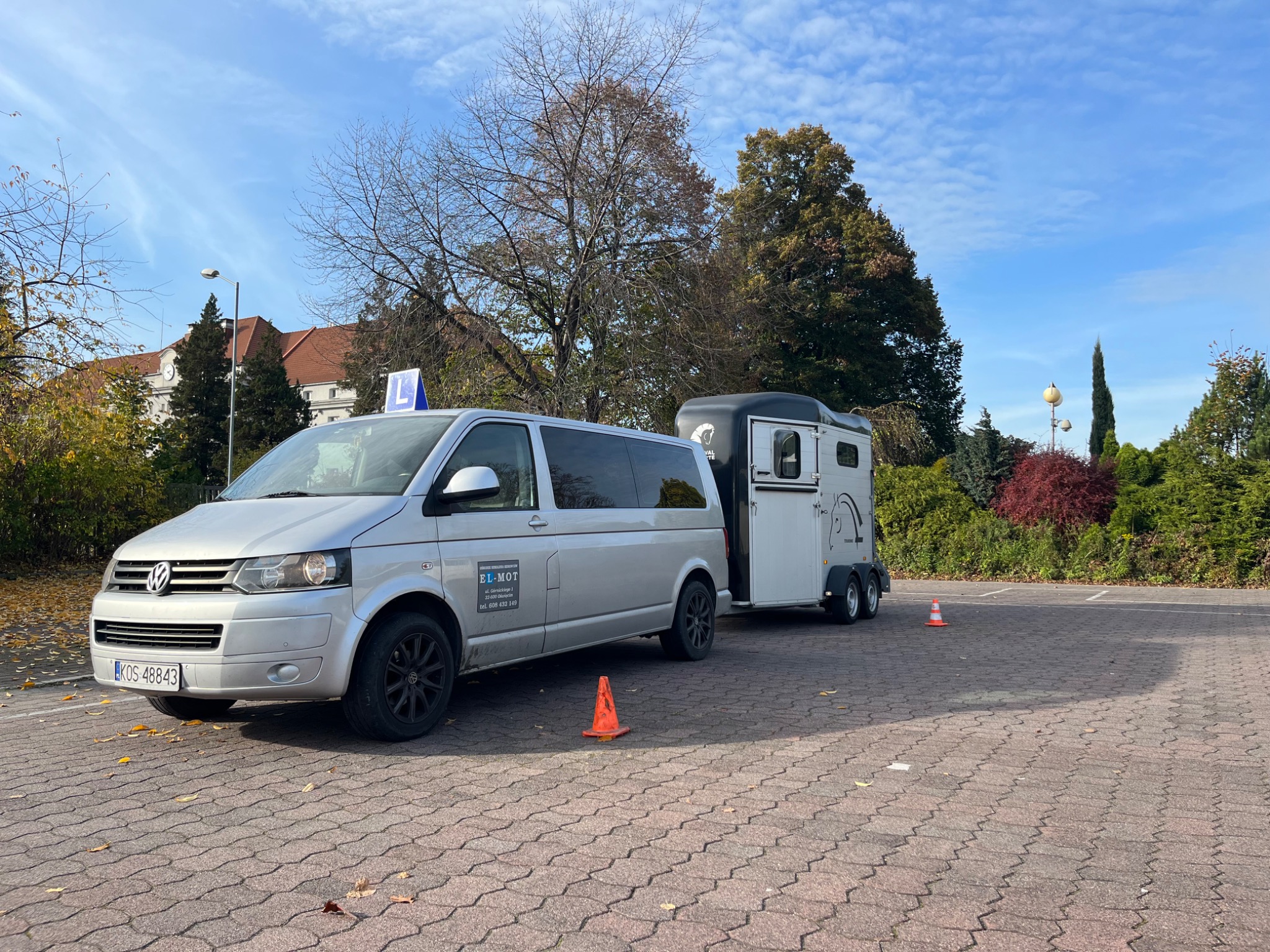 Srebrny van z przyczepą do przewozu koni, oznaczony literą 'L' na dachu, stoi na brukowanym placu, z pomarańczowymi pachołkami w tle drzewa i budynek.