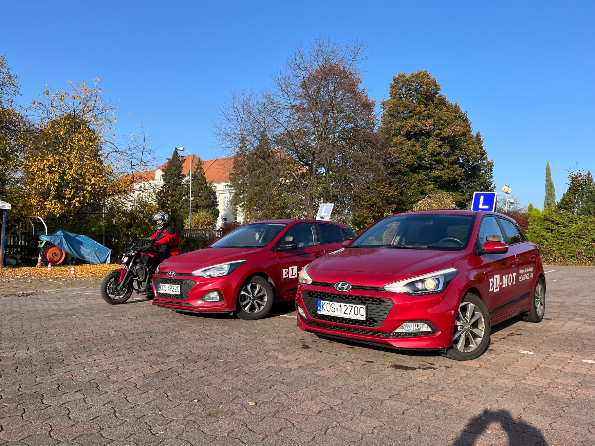 Dwa czerwone samochody z oznaczeniem nauki jazdy na placu manewrowym, obok motocykl z kursantem w kasku i czerwonej kurtce, w tle budynek z czerwonym dachem i drzewa jesienią.