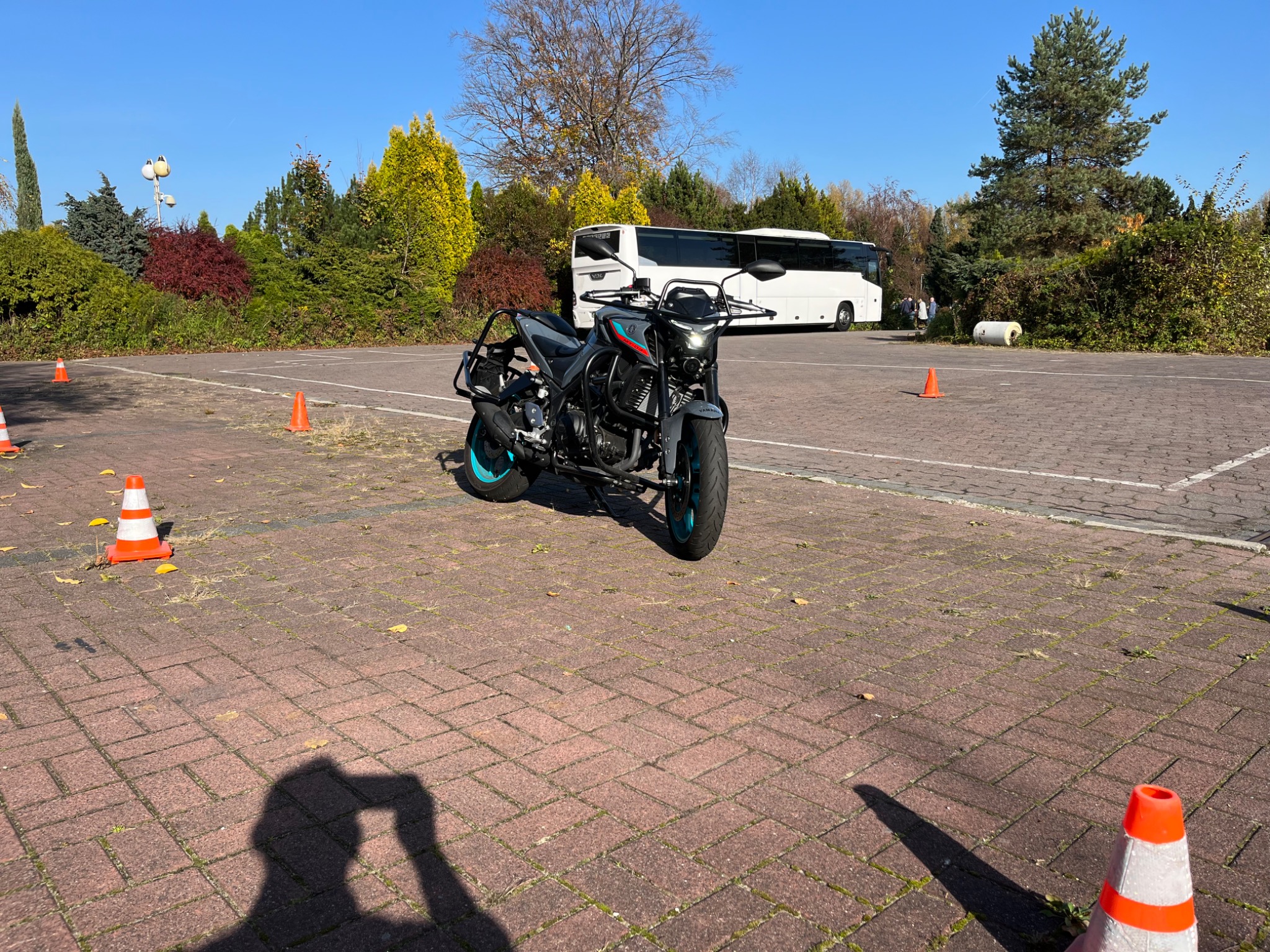 Motocykl na placu manewrowym z pomarańczowymi pachołkami, w tle autobus i zieleń, cień osoby fotografującej w dolnej części kadru.