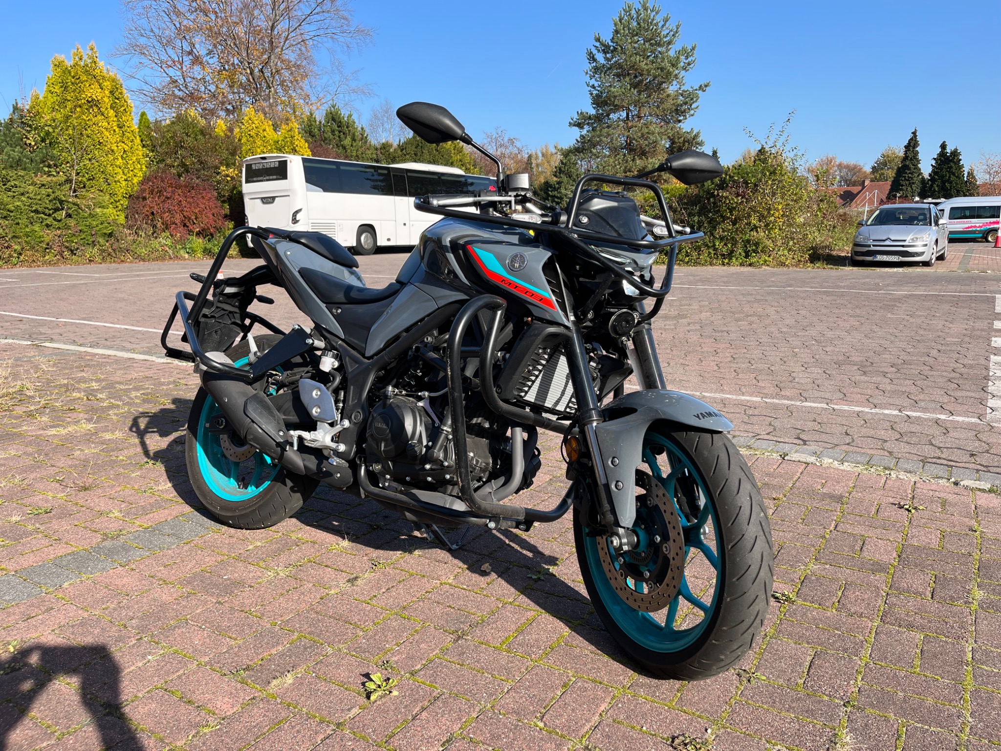 Szary motocykl Yamaha MT-03 z turkusowymi felgami, zaparkowany na brukowanym placu. W tle widoczny biały autokar i inne pojazdy, a także drzewa i krzewy.