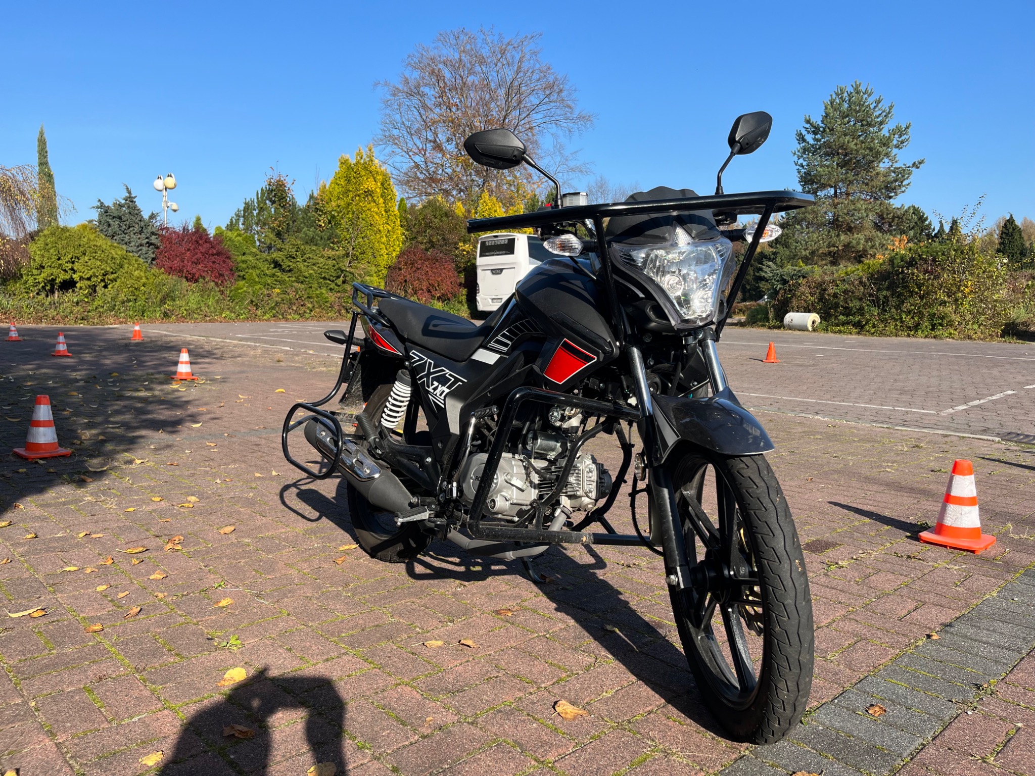 Czarny motocykl treningowy na placu manewrowym w Oświęcimiu, otoczony pomarańczowymi pachołkami, z widocznym białym busem w tle.