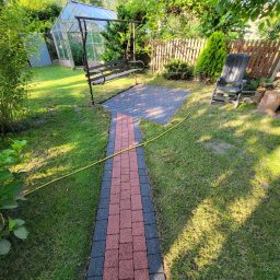 CleanTech - Ułożony z czerwono-czarnej kostki brukowej chodnik prowadzi przez zadbany trawnik w kierunku altany z huśtawką i szklarni, w tle drewniany płot i zieleń drzew.