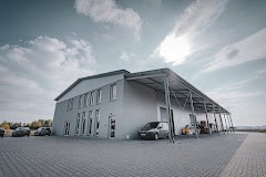 Duży, szary budynek magazynowy z rampą załadunkową i zaparkowanymi samochodami na brukowanym placu, widok z niskiej perspektywy, pochmurne niebo.