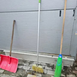 Trzy różne narzędzia do sprzątania: czerwona plastikowa łopata do śniegu, miotła z żółtym włosiem i aluminiowym trzonkiem, oraz zielona miotła z plastikowym włosiem, oparte o szarą ścianę...