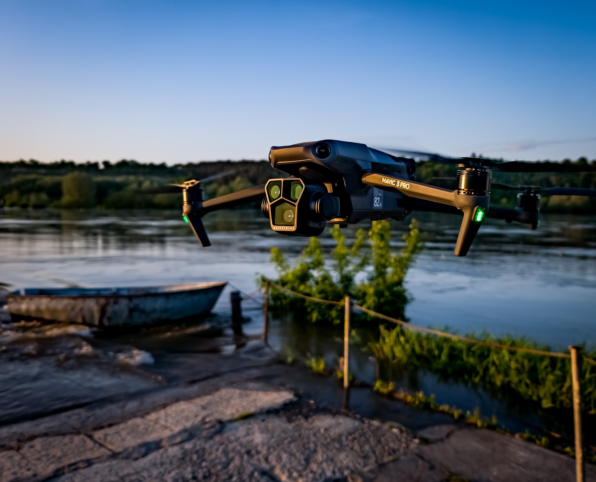 Czarny dron Mavic 3 Pro w locie nad rzeką, w tle zieleń i błękitne niebo o zmierzchu, widoczny fragment betonowego nabrzeża z małą łódką.