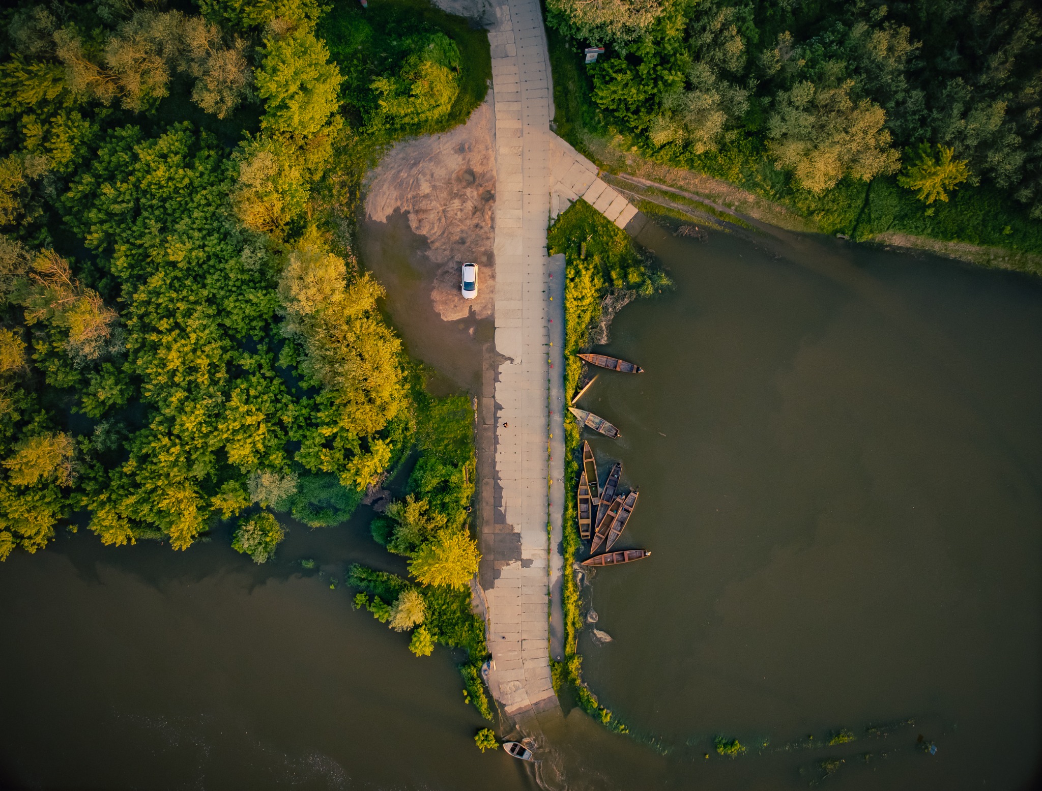 Widok z lotu ptaka na betonowy pomost prowadzący do rzeki z zaparkowanym białym samochodem i zacumowanymi drewnianymi łodziami, otoczony zielenią drzew.