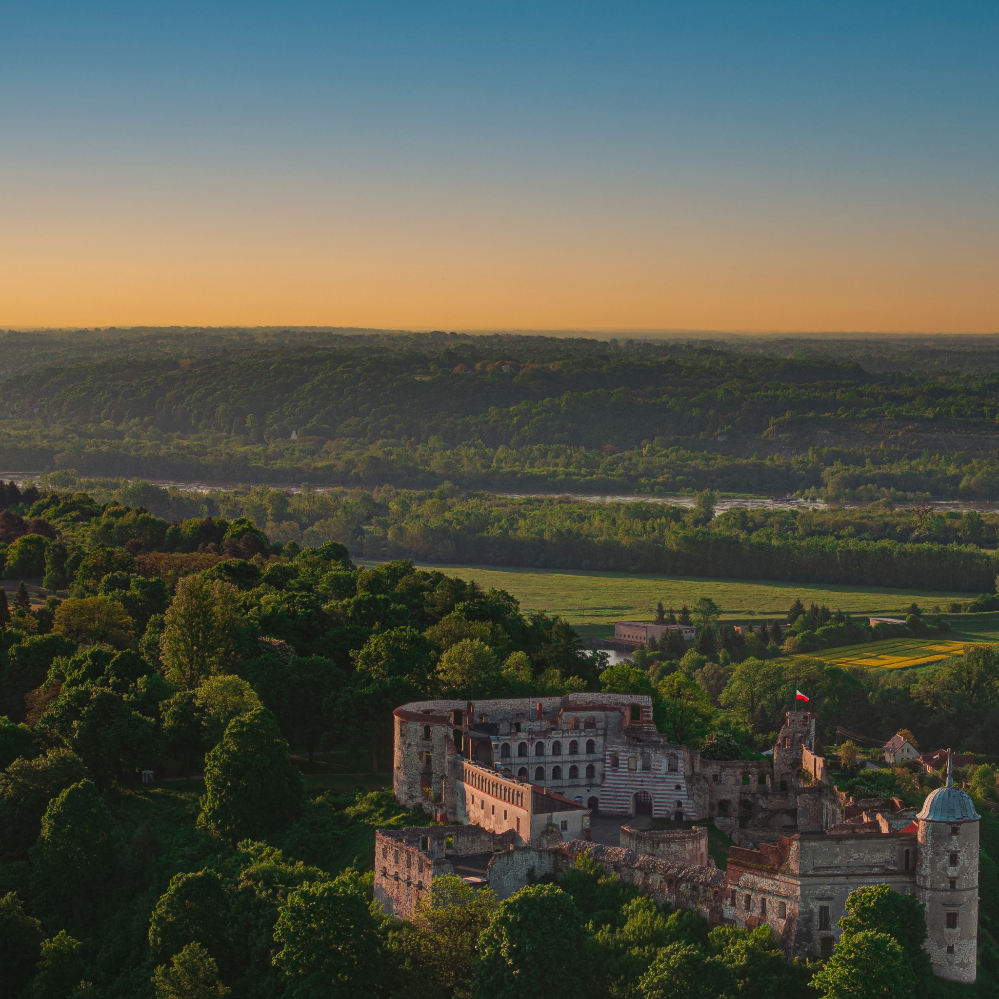 Panorama ruin zamku w Kazimierzu Dolnym, otoczonego zielenią, z widokiem na dolinę Wisły o zachodzie słońca.