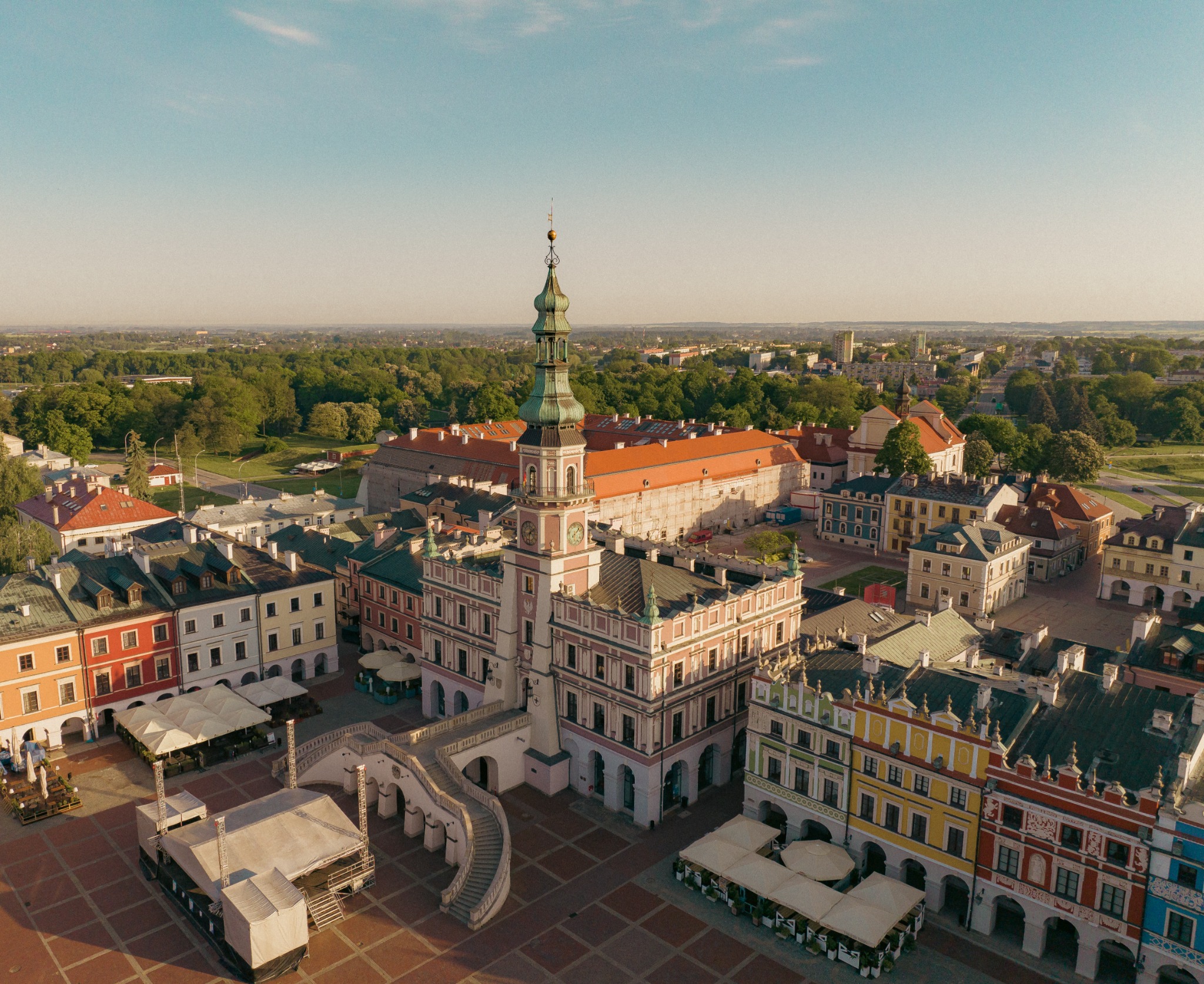 Panorama Zamościa z lotu ptaka, widoczny Rynek Wielki z ratuszem, kolorowe kamienice i scena plenerowa, w tle zieleń drzew i zabudowa miejska w porannym świetle.