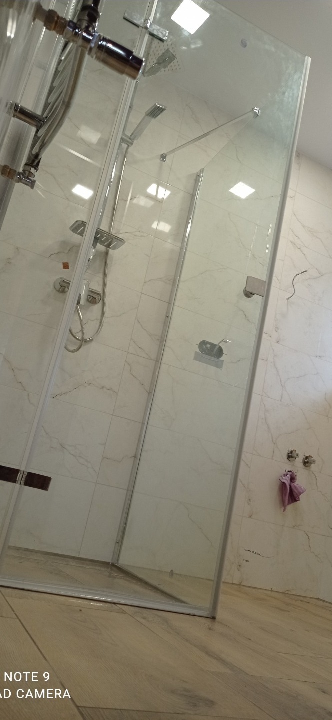 Nowoczesna kabina prysznicowa z chromowaną armaturą i deszczownicą, marmurowe płytki ścienne z delikatnym wzorem żyłkowania, drewnopodobna podłoga, widoczne niedociągnięcia montażowe (przewody)