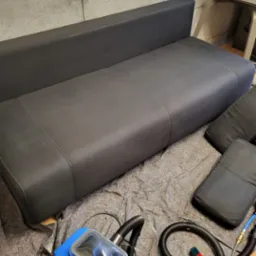 Ciemnoszara kanapa z poduszkami i niebieskim urządzeniem do czyszczenia tapicerki na rozłożonej folii ochronnej.