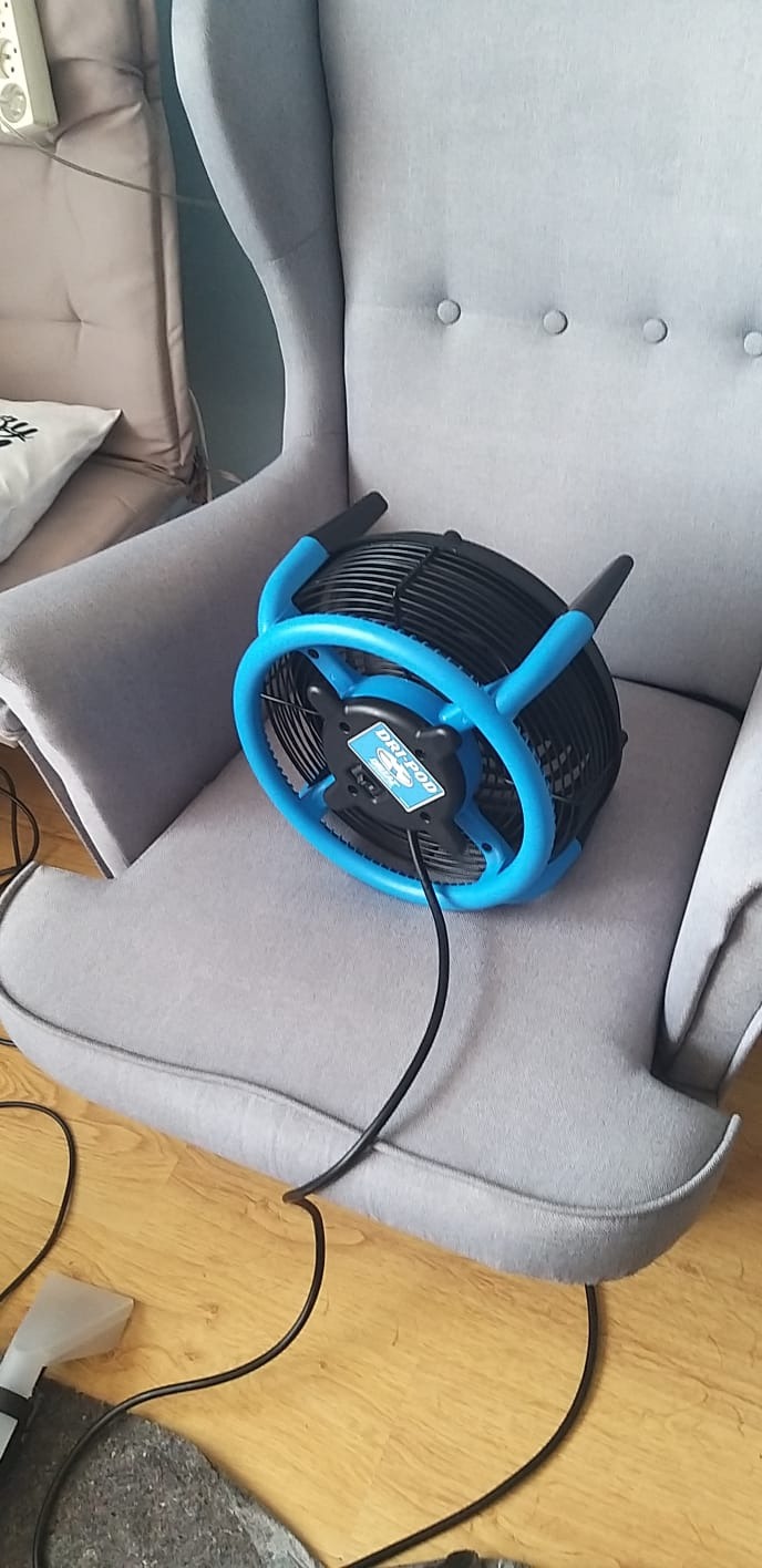 Niebiesko-czarny wentylator przemysłowy marki DRI-POD na szarym fotelu typu uszak, widoczny fragment węża odkurzacza piorącego na drewnianej podłodze.