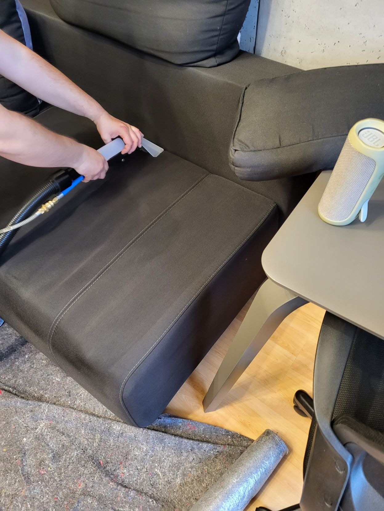 Czyszczenie ciemnoszarej sofy za pomocą urządzenia ekstrakcyjnego; widoczny fragment sofy, ramię osoby obsługującej urządzenie, oraz fragment stolika z głośnikiem JBL.
