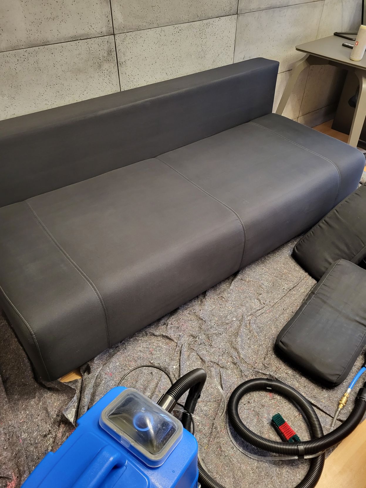 Ciemnoszara kanapa z poduszkami i niebieskim urządzeniem do czyszczenia tapicerki na rozłożonej folii ochronnej.