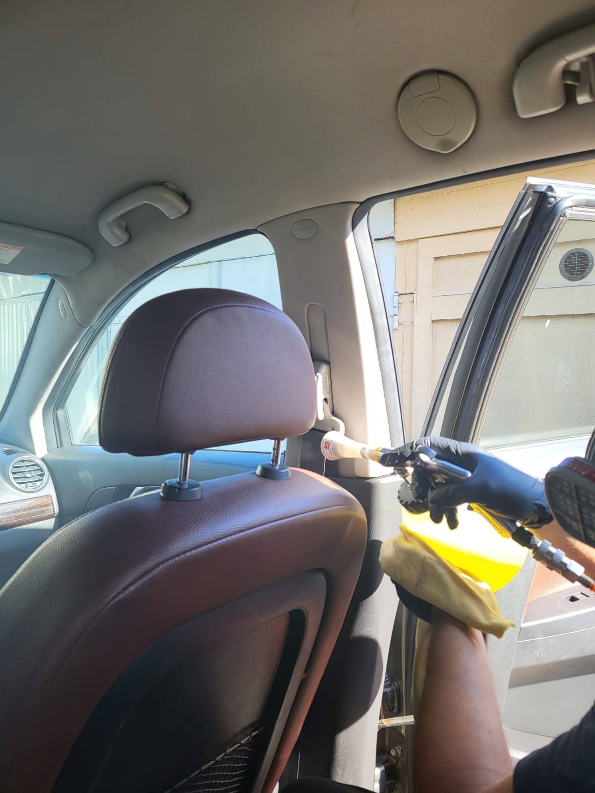Czyszczenie tapicerki samochodowej parownicą, widoczne skórzane siedzenie, dysza parownicy i dłoń w czarnej rękawicy trzymająca ściereczkę.