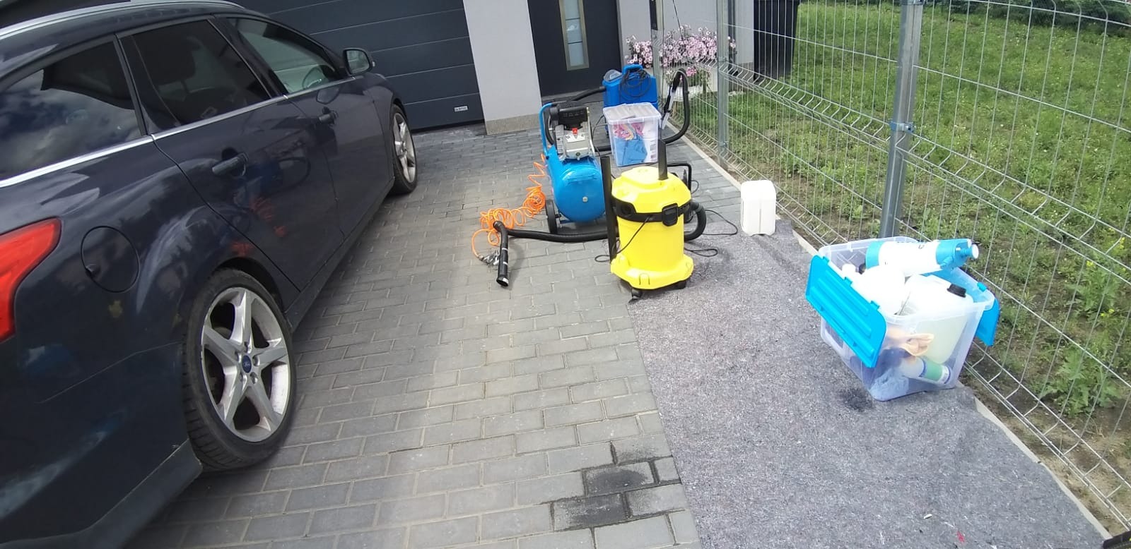 Niebieski samochód zaparkowany obok sprzętu do czyszczenia: żółty odkurzacz przemysłowy, niebieski kompresor i przezroczysty pojemnik z detergentami na kostce brukowej.