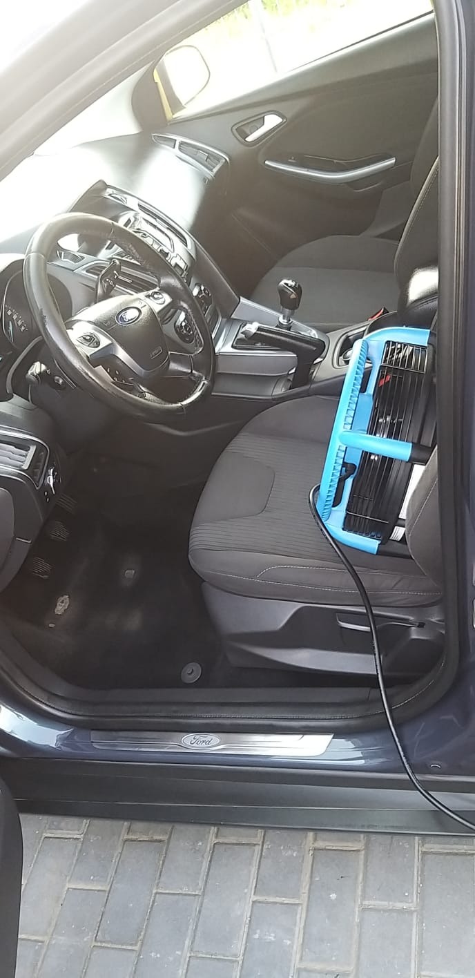 Wnętrze samochodu Ford Focus z szarą tapicerką, na siedzeniu pasażera ustawiona lampa UV z niebieską obudową i czarnym kablem, prawdopodobnie w celu dezynfekcji.