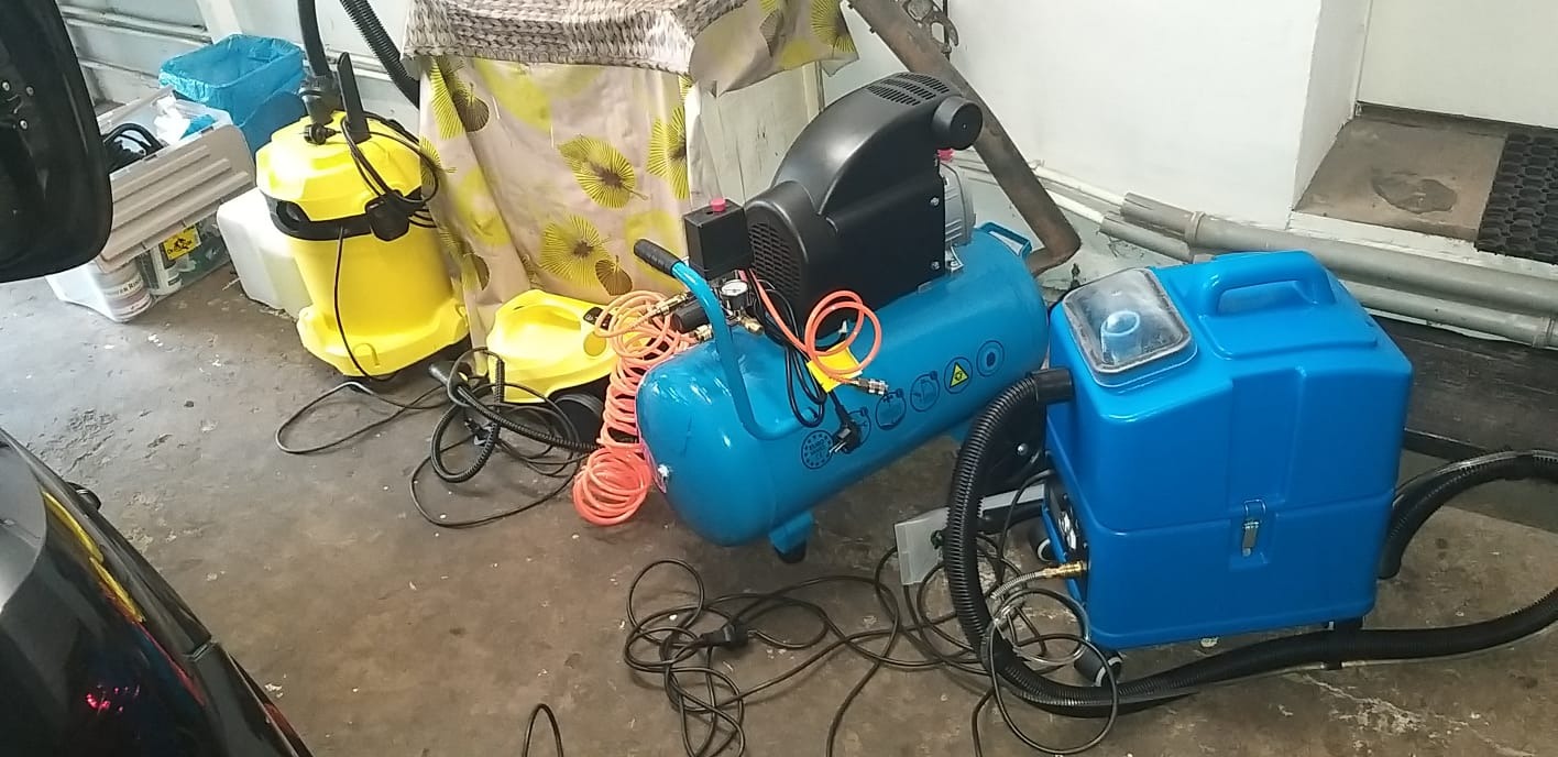 Sprzęt do czyszczenia tapicerki: niebieski ekstraktor do prania, kompresor i żółty odkurzacz przemysłowy na szarym betonowym podłożu, widoczne kable i węże.