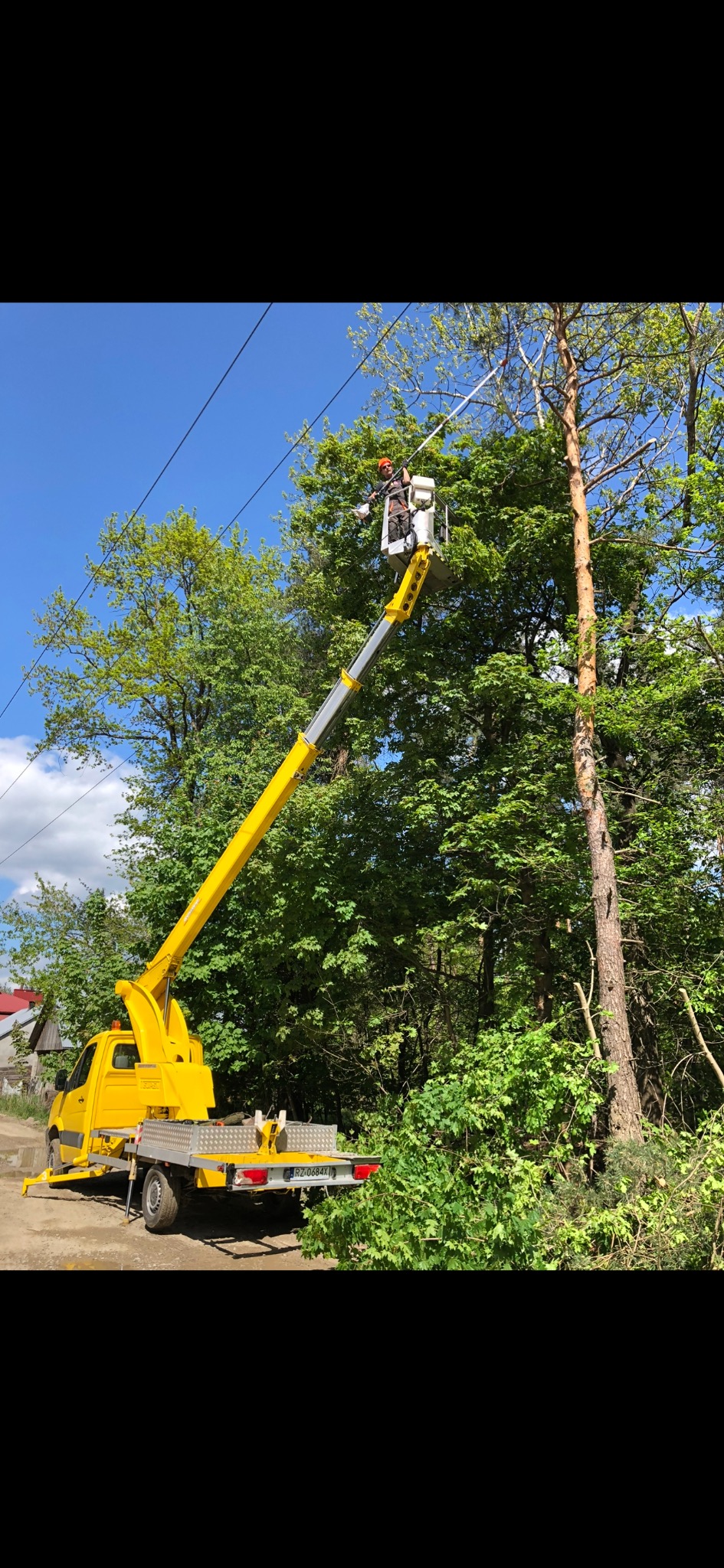 Pracownik na podnośniku koszowym przycina gałęzie drzewa w pobliżu linii energetycznych, widoczny żółty samochód z podnośnikiem i fragment powalonych gałęzi.