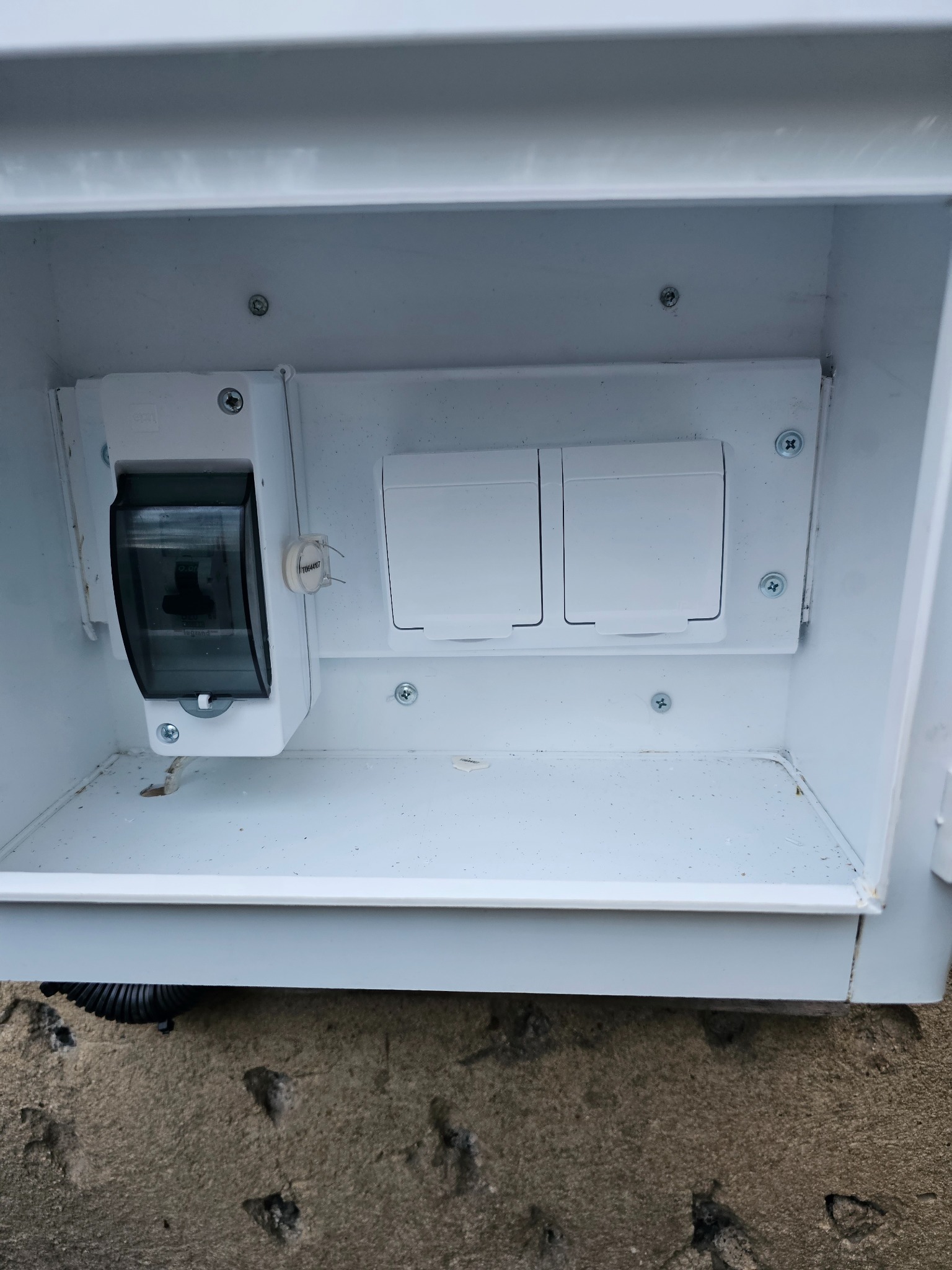 Wnętrze białej skrzynki elektrycznej z zamontowanym wyłącznikiem nadprądowym Legrand z przezroczystą osłoną i dwoma białymi zaślepkami. Widoczne śruby mocujące i fragment peszla u dołu.