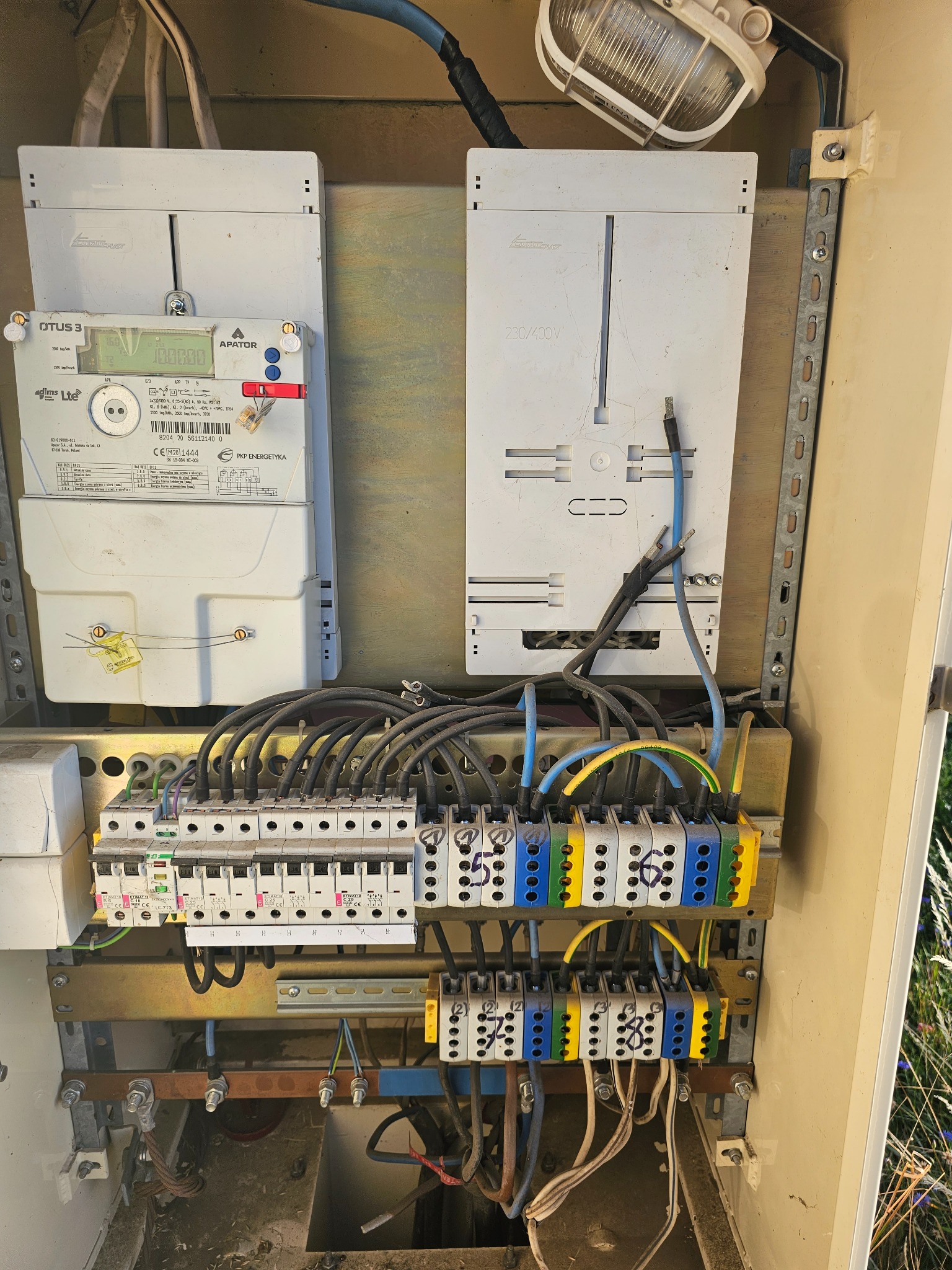 Otwarta skrzynka elektryczna z licznikiem energii, bezpiecznikami i okablowaniem, widoczna lampa oświetleniowa wewnątrz obudowy, instalacja w trakcie przeglądu lub naprawy.