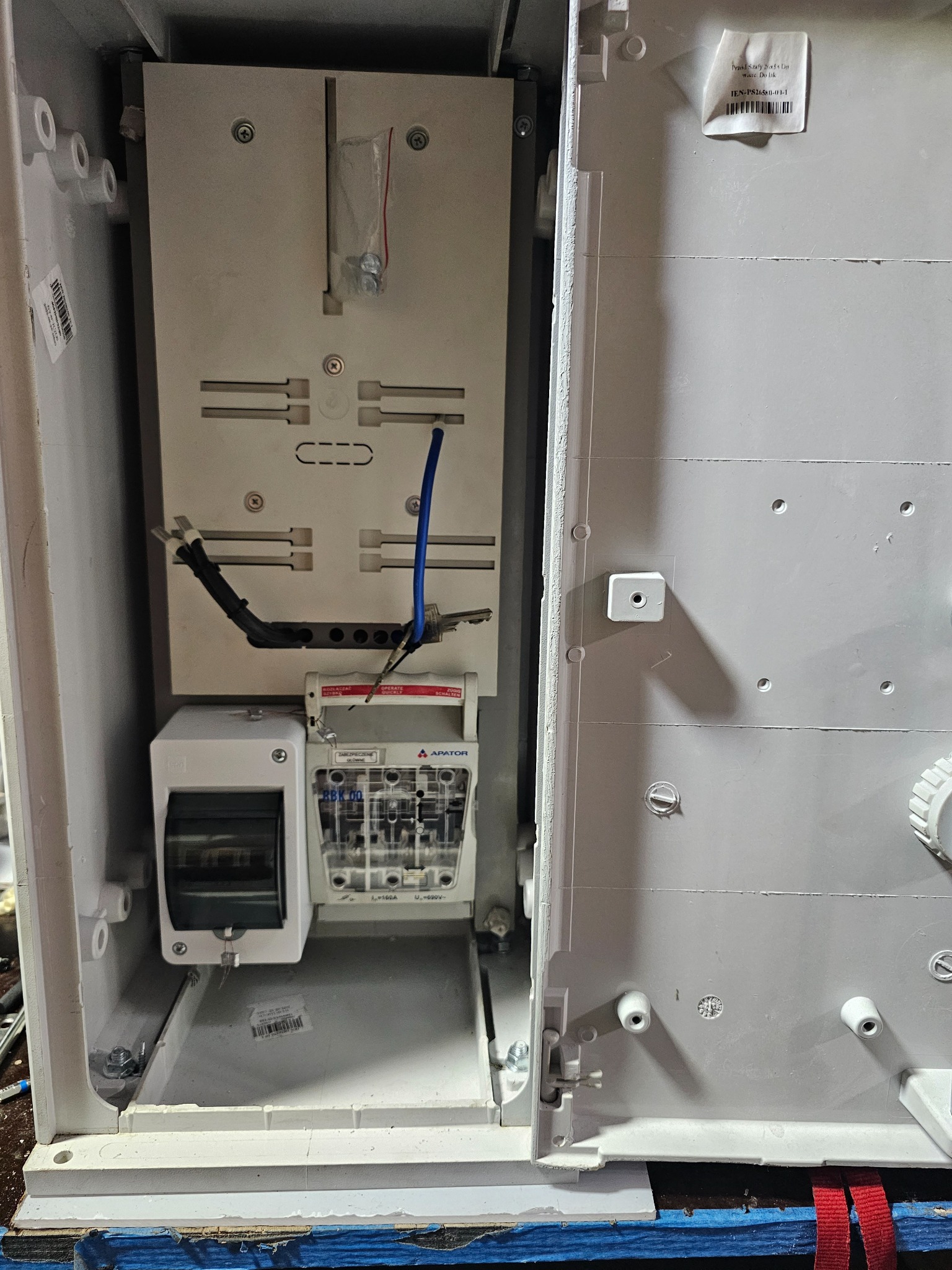 Otwarta, biała skrzynka elektryczna z zamontowanymi bezpiecznikami, wyłącznikami i przewodami, widoczne elementy montażowe i naklejki z oznaczeniami.