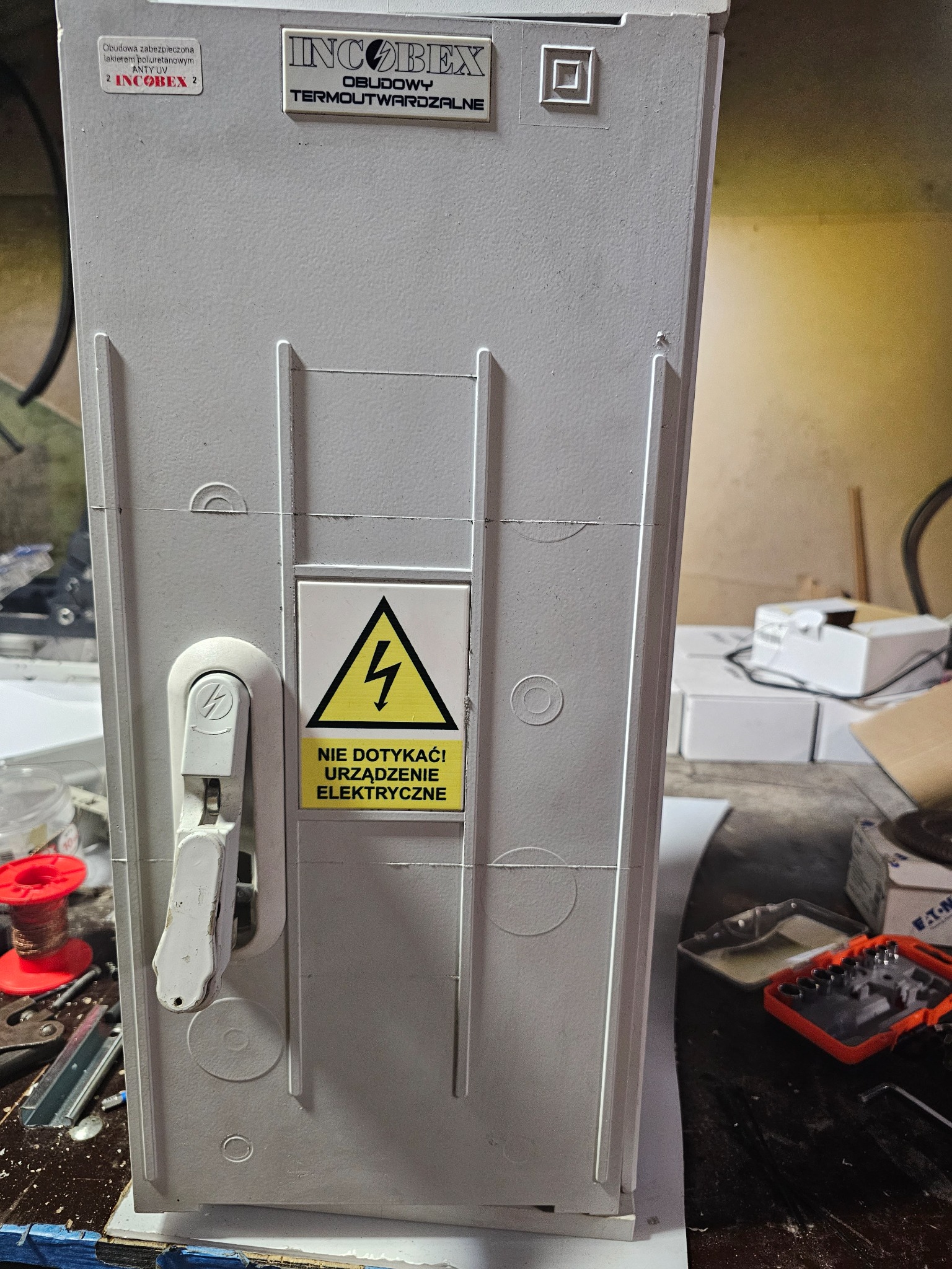 Szara obudowa zabezpieczająca urządzenie elektryczne z napisem ostrzegawczym 'Nie dotykać! Urządzenie elektryczne' sfotografowana w Nekli, widoczne narzędzia i akcesoria elektryczne.