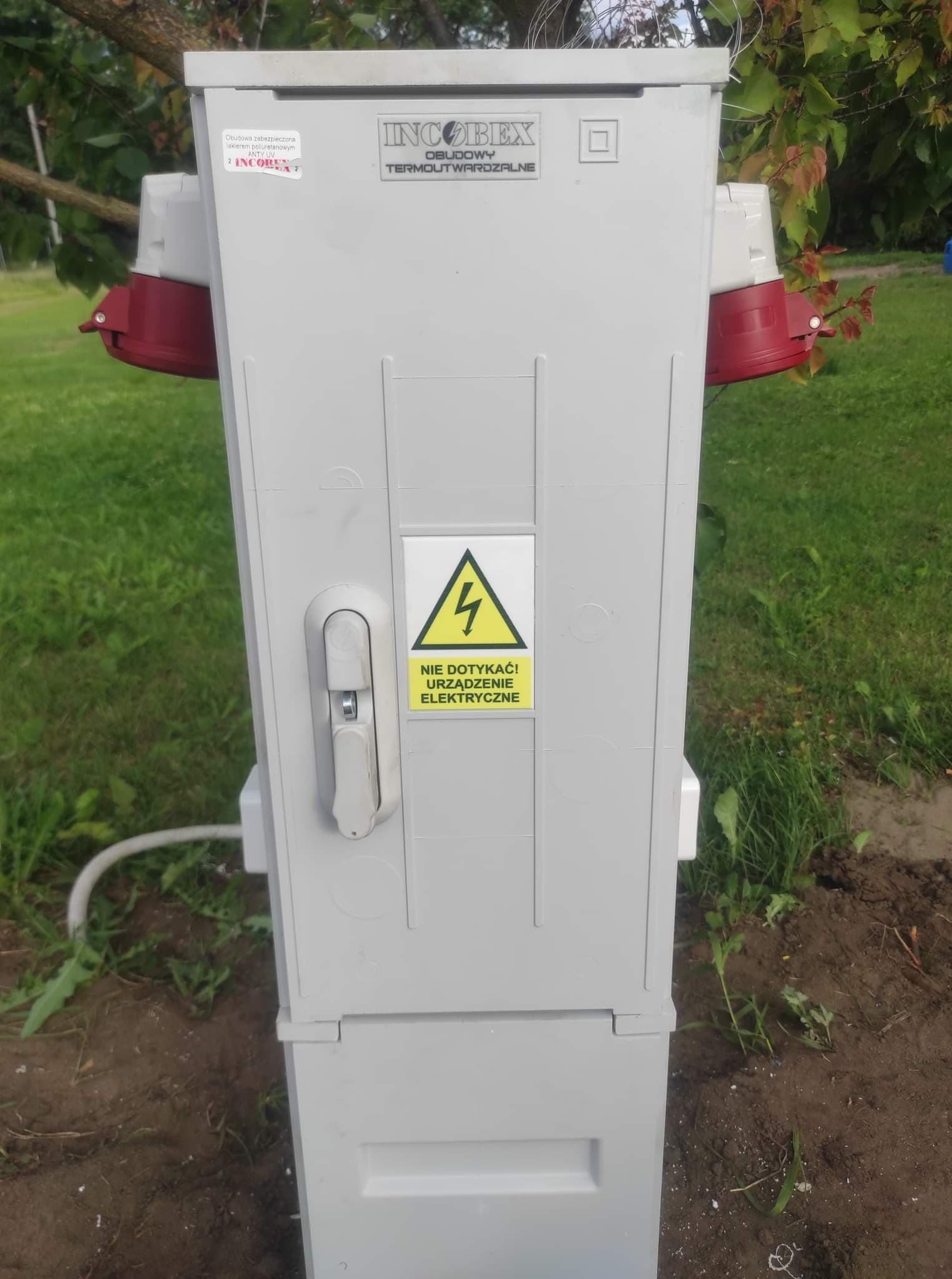 Szara, termoutwardzalna skrzynka elektryczna z gniazdami i ostrzeżeniem 'Nie dotykać! Urządzenie elektryczne', umieszczona na ziemi, z widocznymi elementami instalacji i zielonym tłem trawy i drzew.