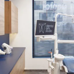 Nowoczesny gabinet stomatologiczny z drewnianymi szafkami, niebieskim blatem i fotelem dentystycznym, wyposażony w lampę zabiegową, monitor z logo 'Dental Arts Studio' oraz aparat RTG.