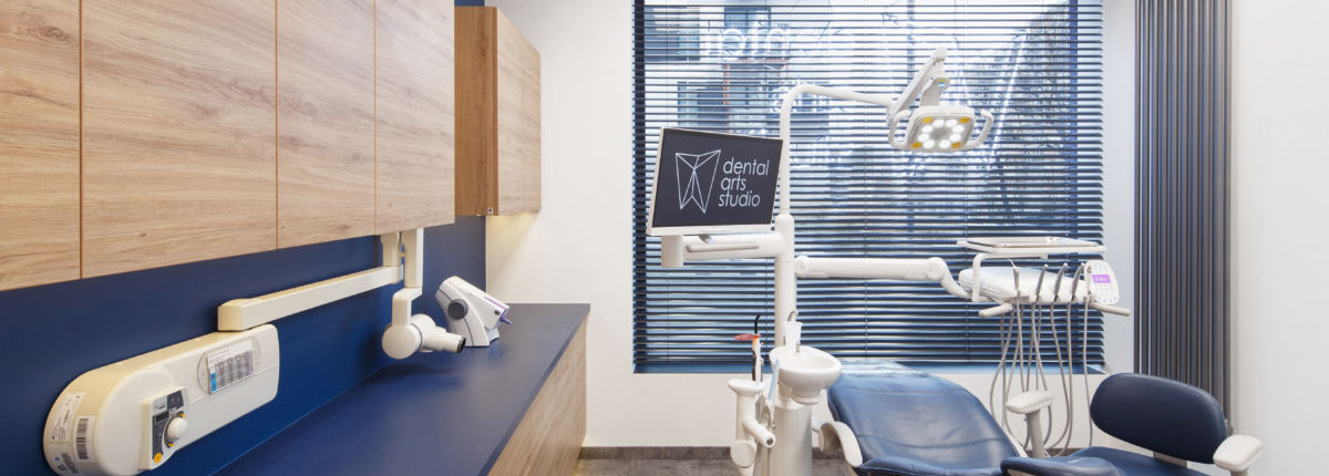 Nowoczesny gabinet stomatologiczny z drewnianymi szafkami, niebieskim blatem i fotelem dentystycznym, wyposażony w lampę zabiegową, monitor z logo 'Dental Arts Studio' oraz aparat RTG.