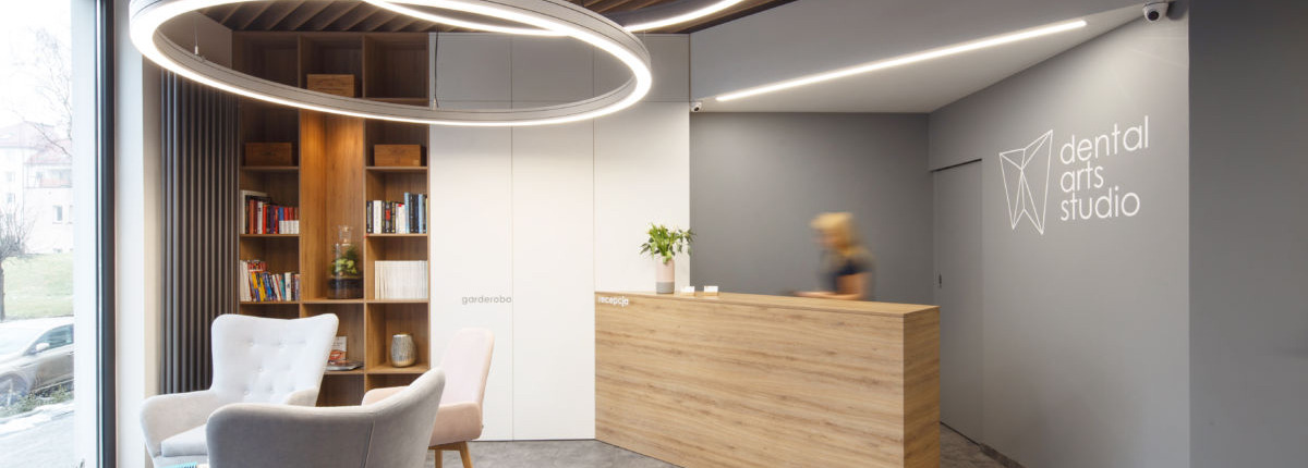 Nowoczesne wnętrze recepcji kliniki z drewnianą ladą, okrągłymi lampami sufitowymi, regałem z książkami i logo 'dental arts studio' na szarej ścianie.